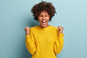 Successful dark skinned female student happy to get scholarship, clenches fists, accomplishes goal, exclaims finally victory, stands amused over blue background. Success, cheer and achievement concept